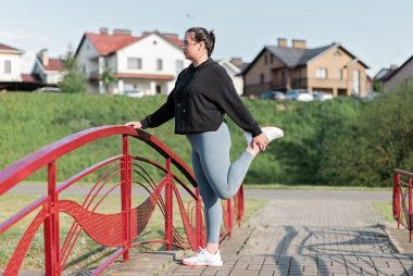 A woman in active wear performs a stretching exercise on a red bridge in a suburban park setting.