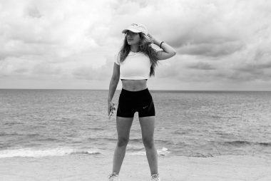 A woman in athletic attire poses near the ocean with a dramatic sky in Natal, Brazil.